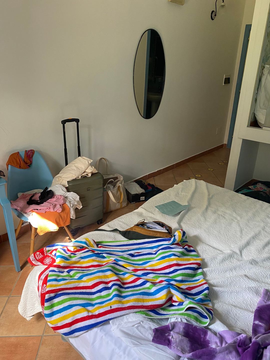 A cluttered hotel room with unpacked luggage, scattered clothing, and personal items on the bed and floor.