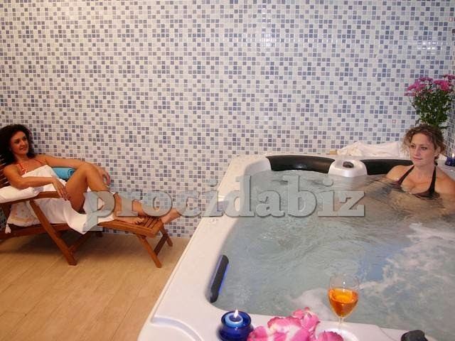 Two guests relax in a hot tub spa facility with one seated in the water and another on a wooden lounge chair beside it.
