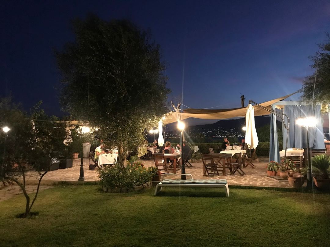 An evening outdoor dining area with illuminated pergolas, tables set for guests, and a scenic hillside view in the background.